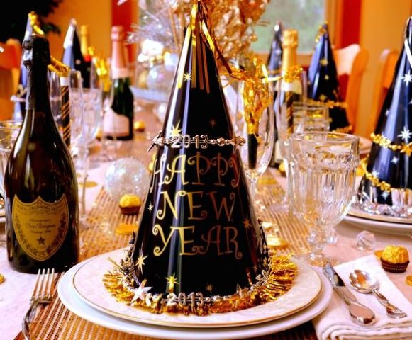 A black and gold NYE party tablescape with a gold sequin table runner, white porcelain and black and gold cone hats plus gold rimmed glasses