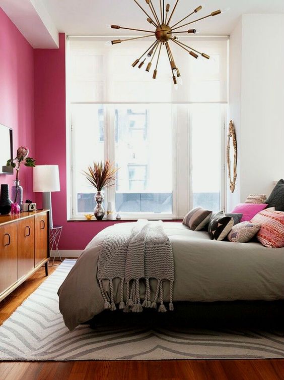A bright and welcoming mid century modern bedroom with a fuchsia statement wall, a rich stained dresser and a brass burst chandelier