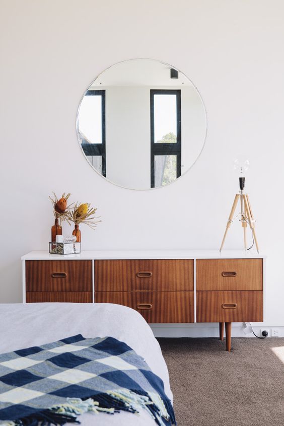 A stylish mid century modern bedroom with a wooden dresser, a round mirror, a large bed and a rug