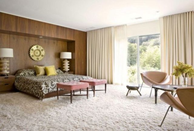 A mid century modern bedroom with a fluffy carpet, a wood covered statement wall, leather chairs and pink stools
