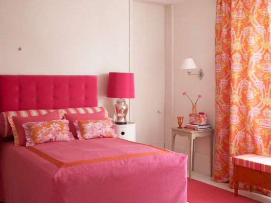 a neutral bedroom with an upholstered bed and pink bedding, a pink table lamp and orange printed curtains, a bright upholstered bench and a table with bold accessories