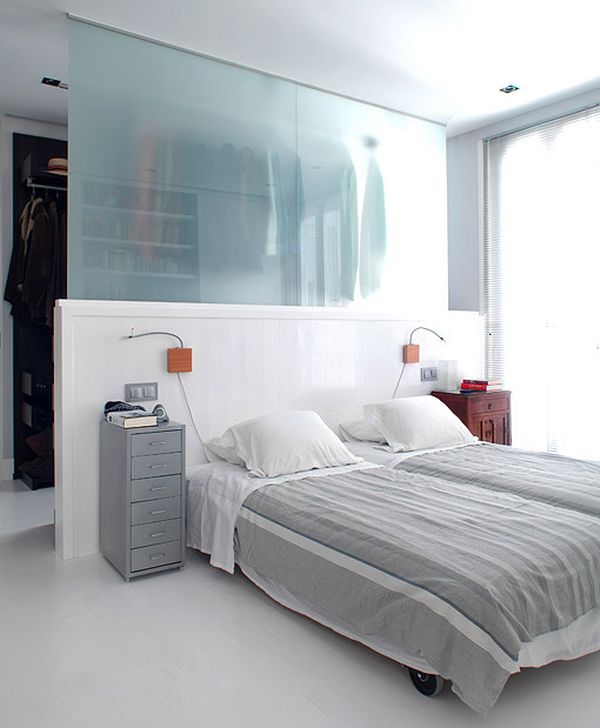 A neutral bedroom with a half wall and a frosted glass wall that divides the bedroom and walk in closet in the space