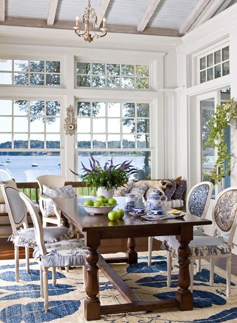 a vintage beach sunroom with a wooden table, blue and white printed chairs with covers, a rug and vintage chandeliers