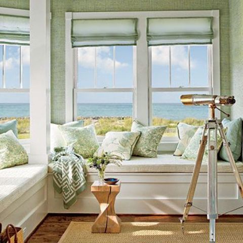 A coastal sunroom with a built in L shaped bench with storage, light green walls, curtains and pillows