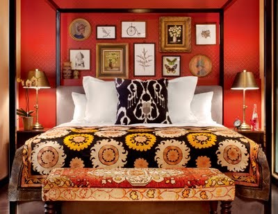 A rust colored bedroom with a canopy bed with bright bedding, a chic gallery wall and metallic table lamps plus an upholstered bench at the foot of the bed