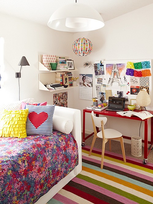 a neutral bedroom made bold mostly with textiles - bedding, a rug, pillows, with a red desk, a colorful gallery wlal and shelves with bright books
