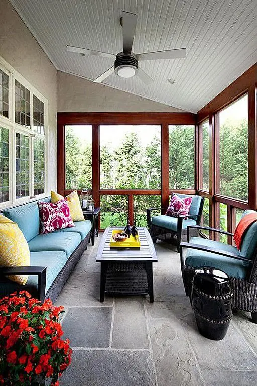 a welcoming and bright screened porch with black wicker furniture with bright upholstery, colorful pillows, a coffee table and bold blooms plus lovely views