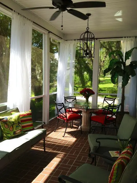 a colorful screened porch with a green sofa and chairs, a round table and red chairs, some curtains for more privacy
