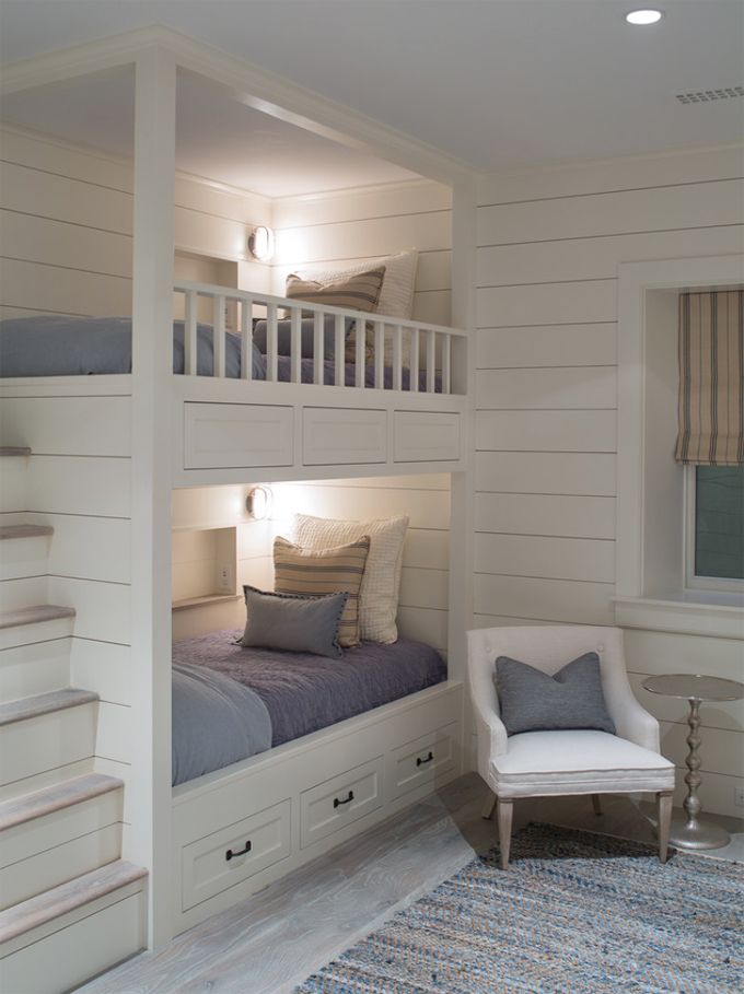 A white coastal bedroom with built in bunk beds, with grey bedding, built in lights and a chair with a grey pillow