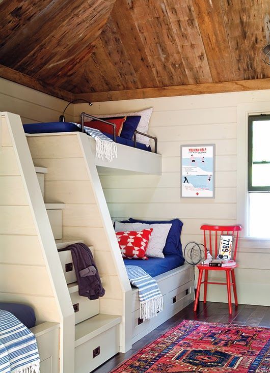 A nautical kids' room with a wooden ceiling, built in bunk beds with bright bedding, a colorful rug and bright artwork is a cool and lovely space