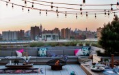 a rooftop summer terrace with a large built-in bench, tables and a fire pit, colorful pillows and string lights over the space