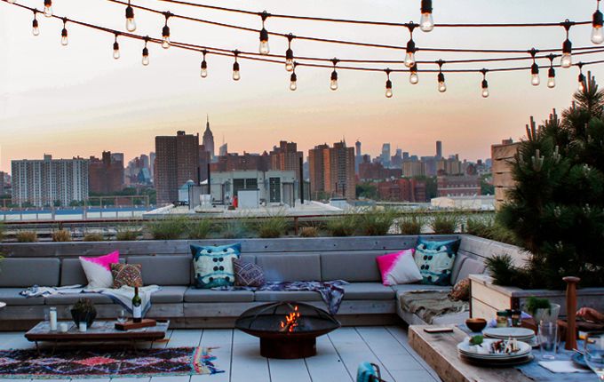 A rooftop summer terrace with a large built in bench, tables and a fire pit, colorful pillows and string lights over the space