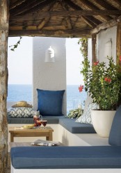 a seaside summer terrace in white, with blue textiles and upholstery, potted blooms and greenery and a cool seaside view