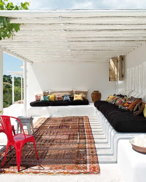 A white summer terrace with a built in bench, dark upholstery and colorful pillows, a boho rug and a cool sea view