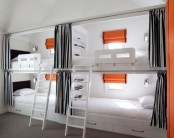 an ultra-modern kids’ room with four built-in bunk beds, with grey curtains, orange touches and seaside inspired sconces