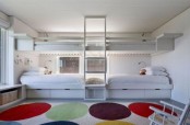 a white kids’ room with built-in bunk beds, a windowsill storage bench, a bright rug, a ladder and some chairs