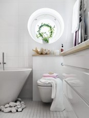 a minimalist white bathroom with a sleek vanity with a neutral stone countertop, an oval tub with pebbles, a round window and greenery