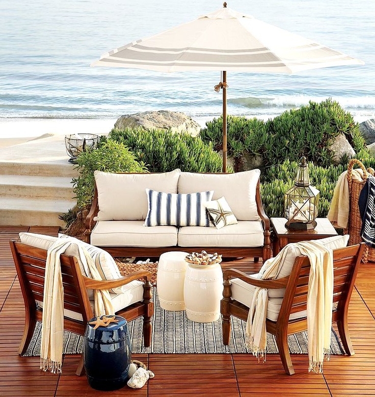 A beach patio with a deck, rich stained wooden furniture with neutral upholstery, candle lanterns and corals and baskets