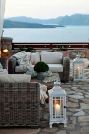 a seaside patio with wicker furniture, neutral upholstery, potted greenery and white candle lanterns features a very lovely view