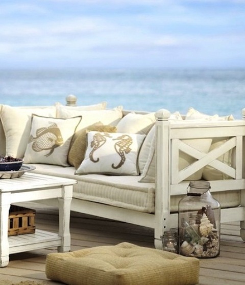 a seaside patio with vintage neutral furniture, neutral upholstery and printed pillows, jars with seashells and crates is very chic