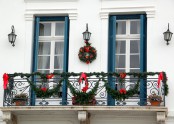greenery garlands, red bows, a greenery wreath with bows and blue shutters give the balcony a bright holiday look