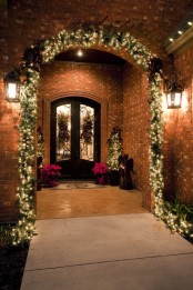 a chic and welcoming Christmas porch with a light fir garland over it, mini lit up trees and hanging plus poinsettia arrangements is very bright and cool