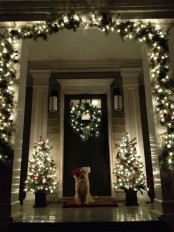 a shiny and festive Christmas porch with a light fir garland over the porch and mini Christmas trees and a wreath looks gorgeous