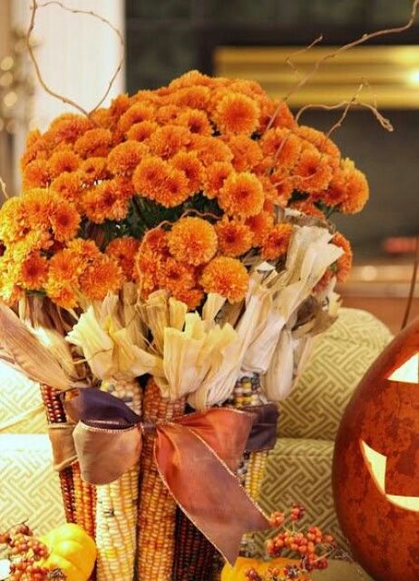 A fall party centerpiece with bright orange blooms covered with corn with husks is a very cool harvest inspired decoration