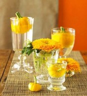 a bright fall party centerpiece of glasses with floating yellow blooms and pumpkins is a whimsy and easy idea