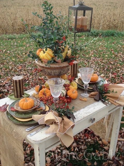A vintage inspired fall table setting with a burlap runner, printed plates, candles, faux pumpkins, berries, a vintage urn with greenery, pumpkins and berries