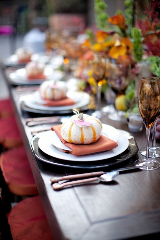 a fall tablescape with dark chargers, glasses, bright napkins and faux pumpkins for each place setting