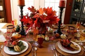 a bold deep red and rust tablescape with an arrangement of fall leaves, candles, napkins, faux pumpkins and gourds plus woven placemats