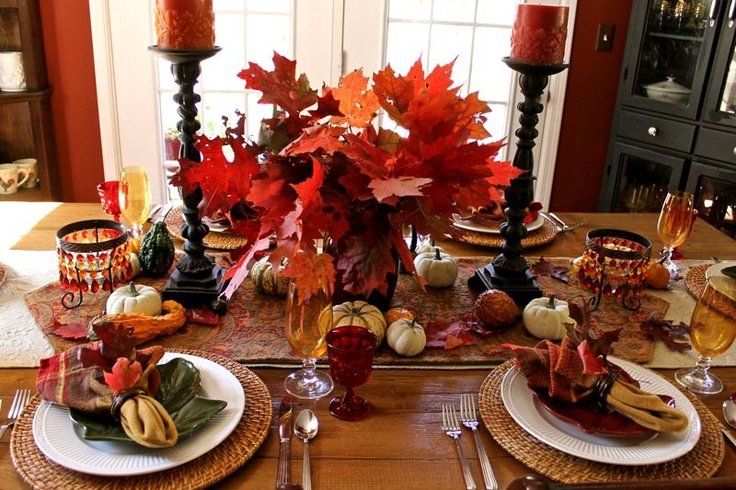 A bold deep red and rust tablescape with an arrangement of fall leaves, candles, napkins, faux pumpkins and gourds plus woven placemats