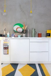 a Scandinavian kitchen with a concrete wall, sleek white cabinets, a colorful geo tile rug and yellow pendant bulbs is all cool