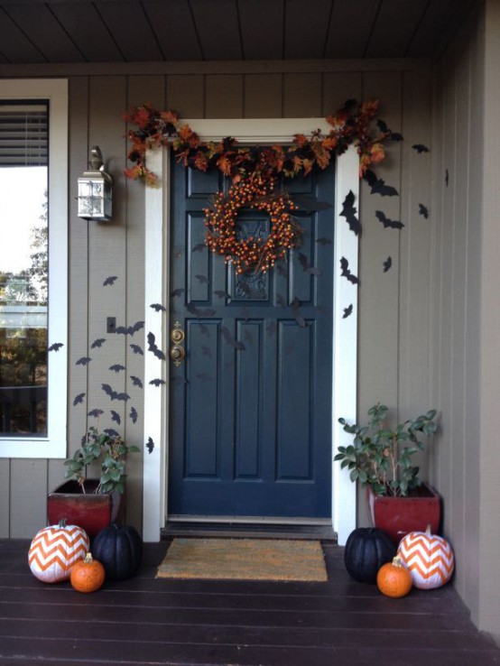 a Halloween front porch with bold pumpkins, plants, black bats attached to the walls and door, a bright leaf garland and a wreath