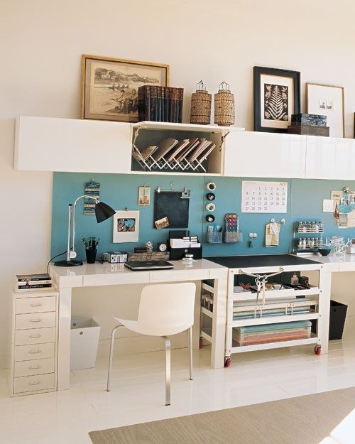 A white shared home office with a large shared desk, a long blue memo board, a sleek wall mounted storage unit is very functional