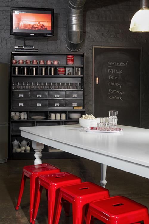 a dark and moody industrial dinign space with black walls, a black storage unit, exposed pipes, a white vintage table and hot red metal chairs