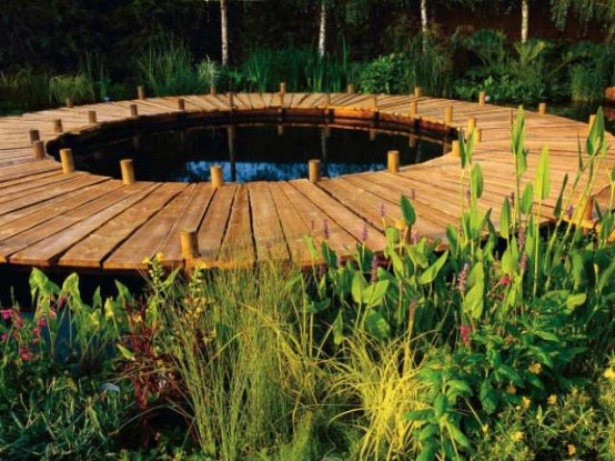 a simple round wooden deck with a pond inside it and lots of greenery around