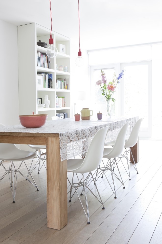 A Nordic dining room with a white bookcase, a light stained wooden dining table, white chairs and pendant bulbs over the space