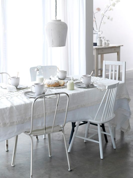 An all white and serene Scandinavian dining room with a table covered with a white tablecloth, mismatching white chairs and a white pendant lamps is super inviting