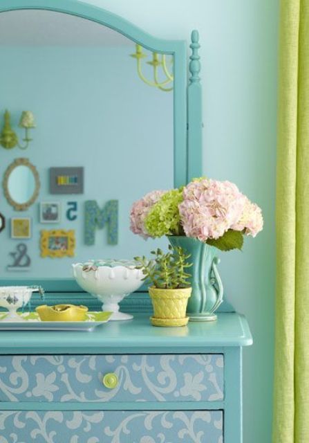 A turquoise room with a turquoise vanity with a mirror and drawers, some pretty decor and mustard colored curtains is wow