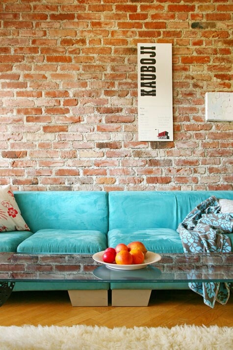 an industrial living room with a brick wall, a turquoise sofa, a glass coffee table, printed textiles and artworks