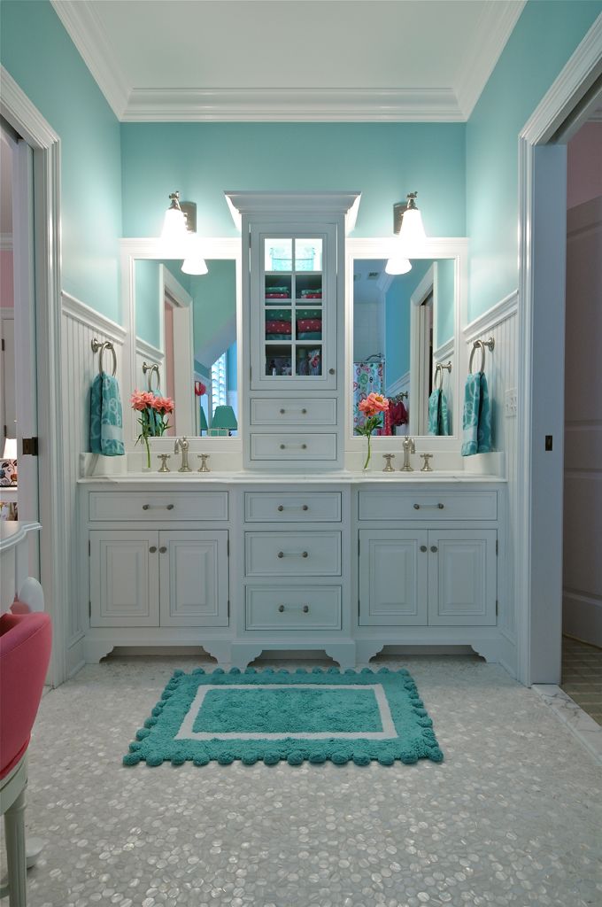 A bathroom with turquoise walls, a white built in double vanity and a storage unit, a turquoise rug and large mirrors and lights