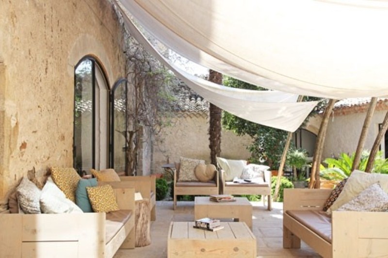 A modern terrace with light colored wooden furniture, printed pillows and potted greenery plus curtains