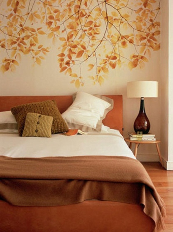 A fall colored bedroom with a leafy wall, an orange bed, muted color pillows, a table lamp with a brown glass base