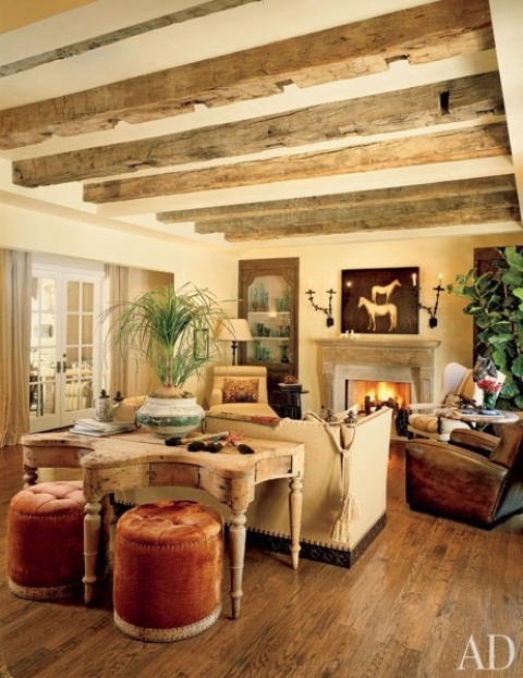 A refined vintage barn living room with wooden beams, a beautiful fireplace, neutral and brown seating furniture, a vintage table, rust colored stools and potted greenery