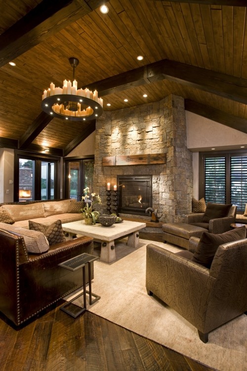 a barn living room with a wooden ceiling and wooden beams, a fireplace clad with stone, neutral leather seating furniture, a low coffee table and a chandelier of candles