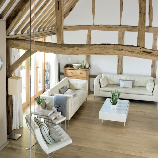 a neutral barn living room with wooden beams and pillars, with neutral seating furniture, white coffee tables, potted greenery and blooms