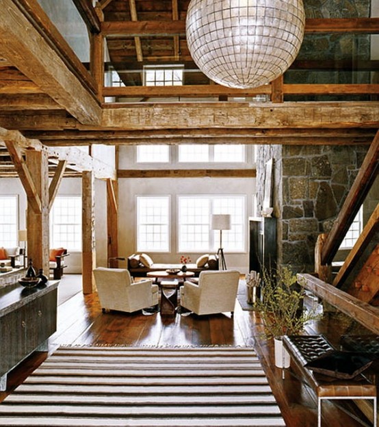 a barn living room with wooden beams and pillas, a fireplace clad with stone, neutral seating furniture and various lamps