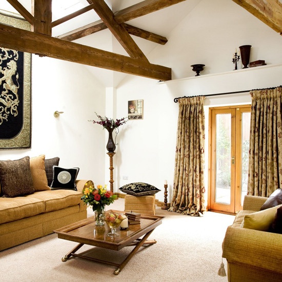 A welcoming barn living room with wooden beams, tan colored seating furniture, bright and printed textiles, a stained wooden table and a coat of arms